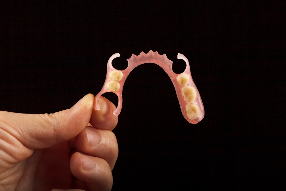 Affordable Partial Dentures - A close-up image of a hand holding a set of affordable partial dentures against a dark background. The partial dentures are designed with a pink, gum-colored base and several artificial teeth, intended to replace missing teeth in a specific section of the mouth. The detailed view highlights the compact and lightweight design of these dentures, which provide a cost-effective solution for restoring dental function and aesthetics without requiring a full set of dentures. The clear focus on the dentures emphasizes their practicality and accessibility for individuals seeking an affordable option to improve their smile and oral health.