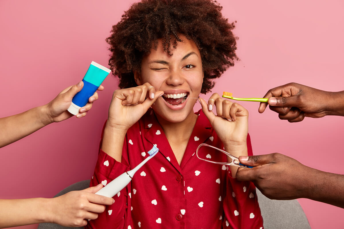 Bad Breath (Halitosis) Remedies - A young woman with curly hair is surrounded by multiple hands offering different oral hygiene tools. She is winking while holding dental floss between her teeth, showcasing the importance of flossing as a remedy for bad breath. The image includes a colorful toothbrush, toothpaste, an electric toothbrush, and a tongue scraper, all aimed at maintaining good oral hygiene and combating halitosis. The woman is wearing a red pajama top with white heart patterns, set against a pink background, emphasizing a cheerful approach to oral care.