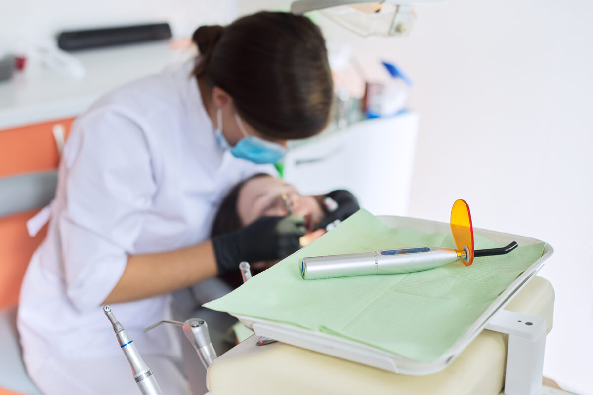 Benefits of Composite Bonding for Teeth - A patient is receiving a composite bonding treatment at a dental clinic. The dentist, dressed in white attire and wearing a face mask, is focused on applying the bonding material to the patient's teeth. In the foreground, a curing light device, equipped with an orange-tinted shield, rests on a green sterile cloth, ready to be used to harden the composite material. The environment is clean and organized, with dental tools neatly arranged, highlighting the professional and hygienic setting. The composite bonding procedure offers benefits such as repairing chipped teeth, enhancing aesthetics, and improving overall dental health.