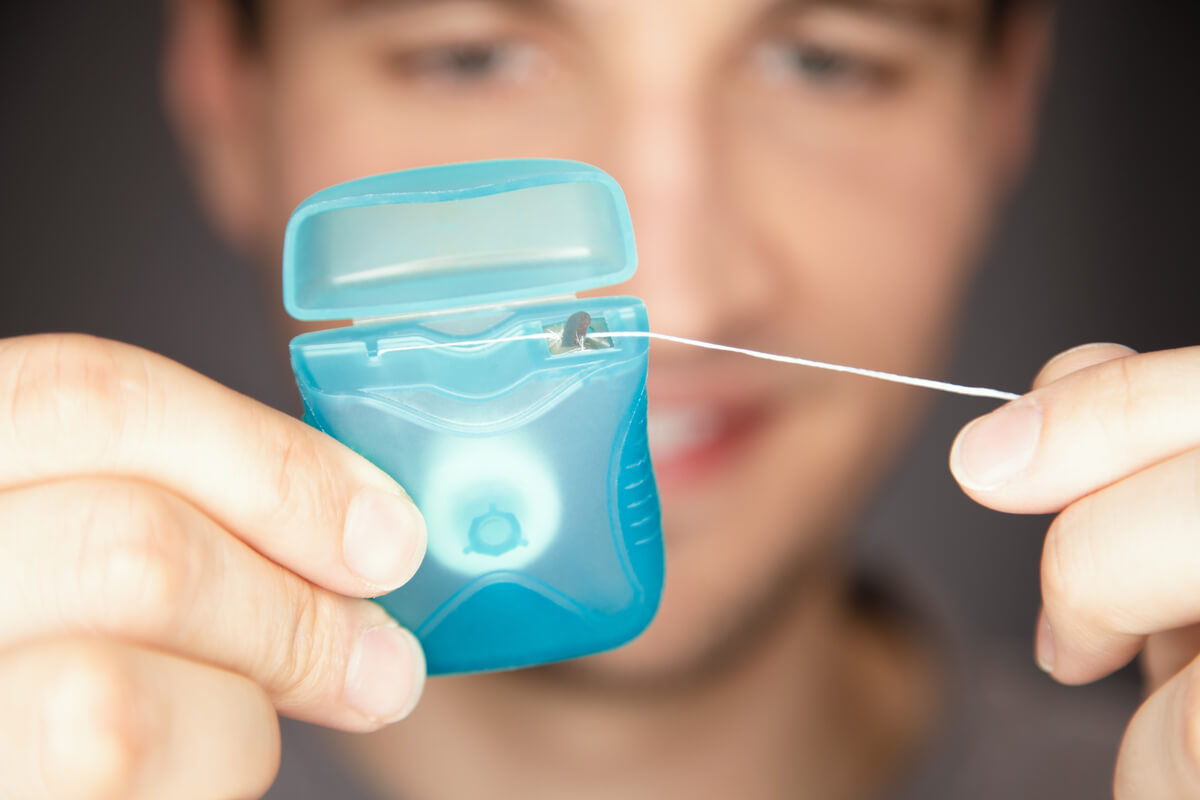 Best Way to Floss Your Teeth - A close-up image of a person holding a container of dental floss, pulling out a strand to use. The focus is on the floss and the hands, emphasizing the preparation for proper flossing. The person’s smile in the blurred background suggests the importance of regular flossing for maintaining healthy teeth and gums. The image highlights the best way to floss your teeth by showing the correct technique for extracting and using dental floss. The clear and detailed view of the floss container reinforces the significance of using the right tools for effective oral hygiene.