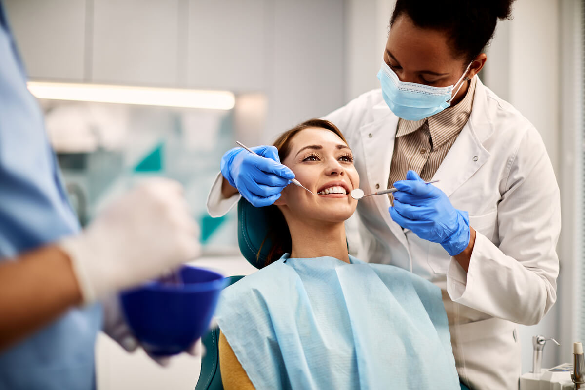 Beyond Bleaching - A patient is undergoing a dental check-up in a modern dental clinic. The dentist, wearing a white coat, face mask, and blue gloves, is using a dental mirror and an explorer tool to examine the patient's teeth. The patient is sitting in a dental chair, smiling and appearing relaxed, covered with a protective blue bib. The setting includes standard dental equipment and tools, indicating a focus on oral hygiene and preventive care. This scene highlights the importance of routine dental visits beyond just teeth whitening procedures to ensure comprehensive oral health.