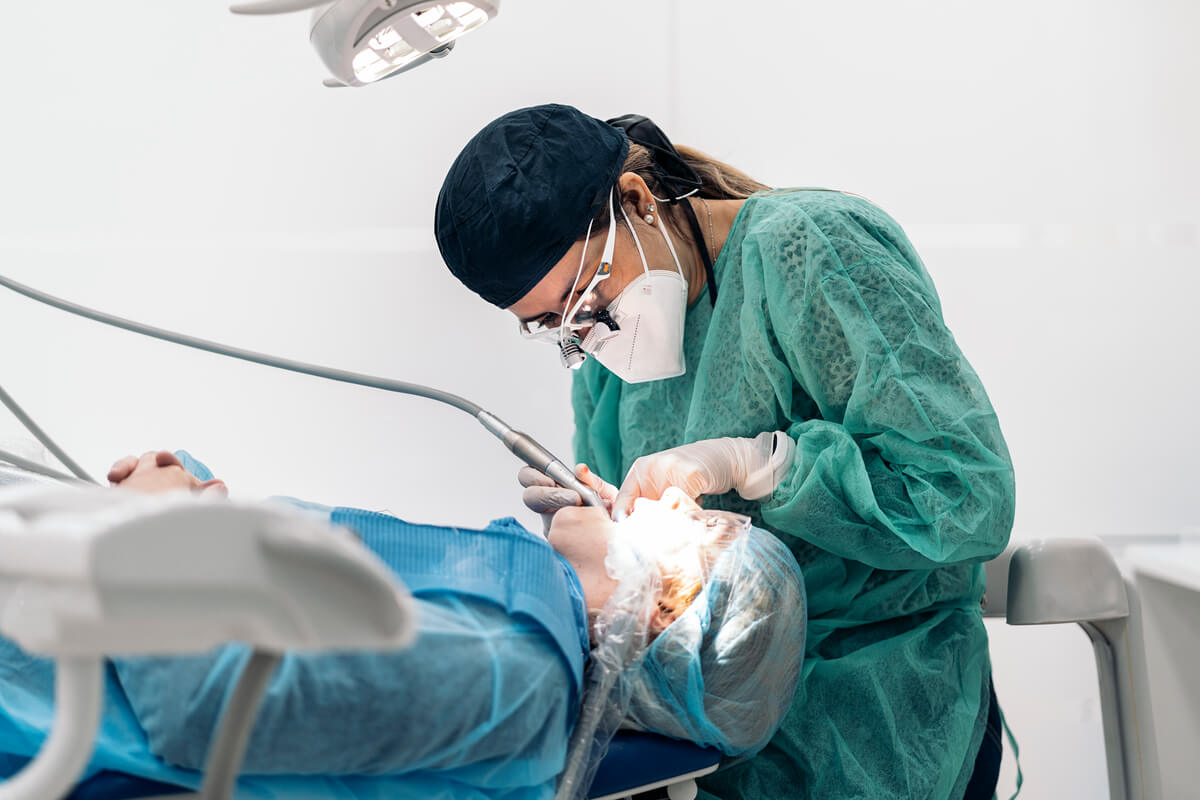 Bone Grafting for Dental Implants - A dental surgeon, dressed in sterile green surgical attire and wearing a mask, performs a bone grafting procedure in preparation for dental implants. The surgeon is focused on the task, using specialized dental instruments under the bright overhead light in a clean, clinical setting. The patient, lying in the dental chair, is draped in surgical blue coverings, with additional protective gear including a cap and face drape, indicating a sterile environment. The surgeon is using magnifying loupes to ensure precision during the bone grafting process, which is crucial for providing a stable foundation for future dental implants.