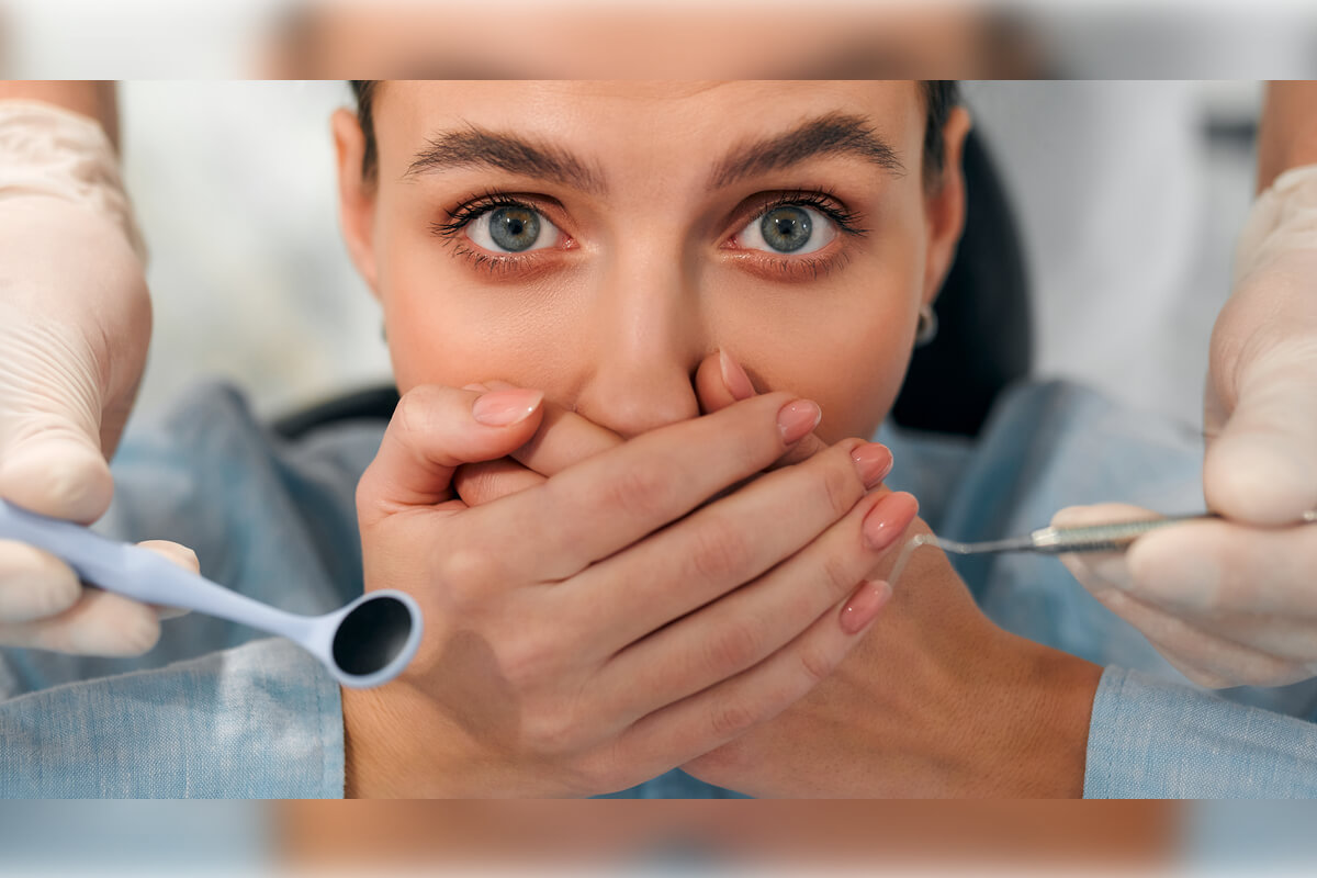 Causes of Bad Breath (Halitosis) - A woman is seated in a dental chair, visibly anxious and covering her mouth with both hands. A dentist, partially visible, is holding dental instruments, including a mirror and a scaler, close to the woman's face. The setting is a dental clinic, where the patient is likely seeking treatment for halitosis, a condition commonly caused by poor oral hygiene, gum disease, or other dental issues. The woman’s wide-eyed expression highlights her fear or concern about the dental examination.