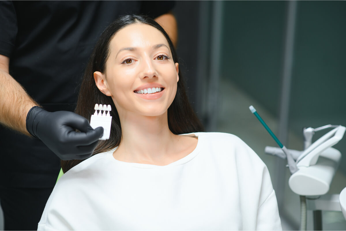 Composite Bonding - A female patient is sitting in a dental chair, smiling while a dental professional holds up a shade guide to match the color of her teeth. The dentist, wearing black gloves, is in the process of selecting the appropriate composite resin color for the bonding procedure. The background shows typical dental equipment, emphasizing the clinical setting. This composite bonding procedure aims to correct imperfections, improve tooth appearance, and provide a natural-looking smile. The patient’s expression and the use of the shade guide highlight the meticulous care involved in achieving aesthetic dental results.