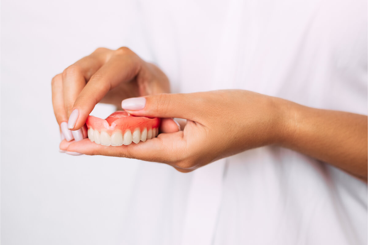 Custom Dentures - A person is holding a set of custom dentures in their hands. The dentures, which include artificial teeth mounted on a pink gum-like base, are carefully being inspected or adjusted. The individual appears to be a dental professional, possibly in a clinic setting, as the background is plain and uncluttered. The custom dentures are designed to replace missing teeth, improve bite function, and enhance the patient's smile, providing a personalized dental solution.