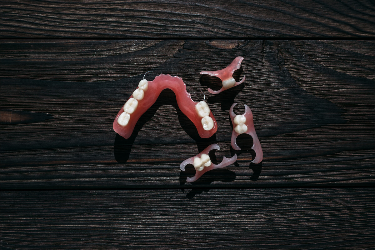 Custom-Made Partial Dentures - A set of custom-made partial dentures is displayed on a dark wooden surface. The dentures consist of several pieces, each designed to replace missing teeth in specific areas of the mouth. The pink gum-colored base and white artificial teeth closely mimic the natural appearance of gums and teeth, offering a personalized fit for the patient. The shadows and natural light emphasize the craftsmanship and detail involved in creating these partial dentures, which provide a practical and tailored solution for restoring dental function and aesthetics in a way that complements the patient’s existing teeth.