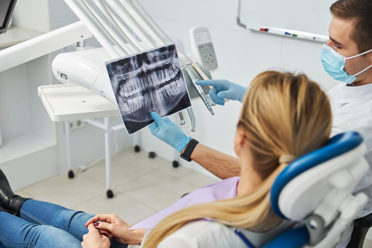 Dental Imaging X-Rays - A dentist, wearing a mask and blue gloves, is showing a dental X-ray image to a patient seated in a dental chair. The X-ray is a detailed panoramic view, capturing the entire set of the patient's teeth and jaw structure. The dentist is pointing to specific areas on the X-ray, likely discussing the patient's dental health or treatment plan. The setting is a modern dental clinic, with clean, white surroundings and advanced dental equipment visible, emphasizing the role of Dental Imaging X-Rays in accurate diagnosis and personalized dental care.