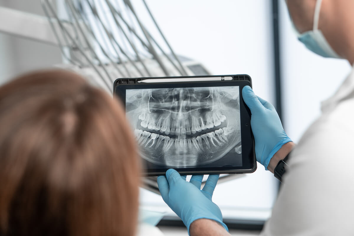Digital Dental X-Rays - A dentist is holding a tablet displaying a clear digital dental X-ray of a patient's teeth and jaw. The dentist, wearing blue gloves and a mask, is showing the detailed image to the patient, who is visible from behind, seated in a dental chair. The digital X-ray highlights the teeth, roots, and bone structure, showcasing the precision and clarity of modern dental imaging technology. The clinical setting is equipped with advanced dental tools, emphasizing the role of digital X-rays in accurate diagnosis and effective dental treatment planning.