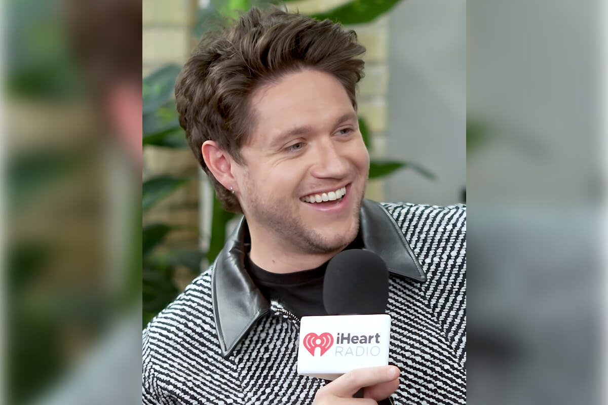 5 Celebrity Smiles - A popular musician, known for his infectious personality, is seen laughing joyfully during an interview session with iHeartRadio. He holds a microphone with the recognizable iHeartRadio logo, indicating a live or recorded discussion. His hair is casually styled, complementing his laid-back fashion choice of a striped sweater with a sleek leather collar. The artist’s smile reveals a genuine happiness, making him relatable and endearing to his fans. The backdrop of green foliage provides a fresh, vibrant feel to the interview setting, emphasizing a relaxed atmosphere often found in entertainment talk shows and radio interviews. This scene captures the essence of how celebrities use their authentic smiles to connect with their audience and convey warmth and positivity.