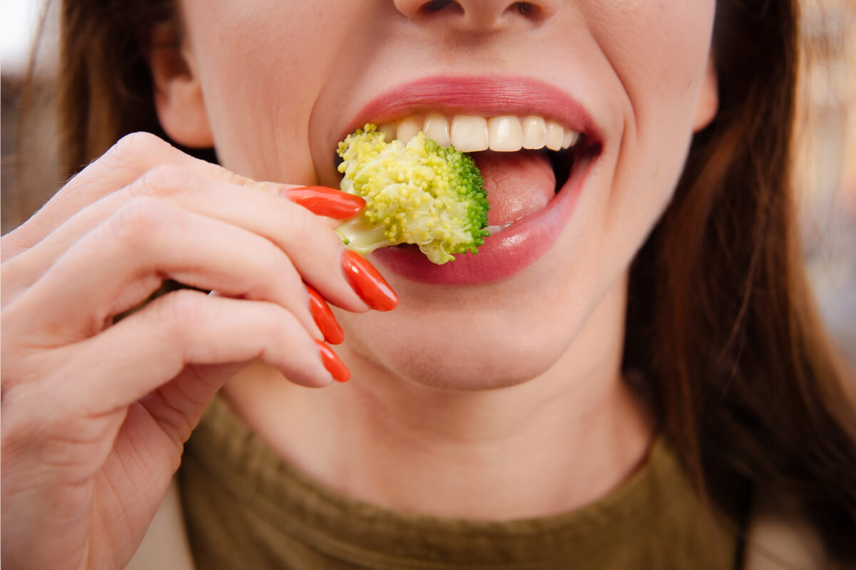 Nutrition and Oral Health - A close-up of a woman taking a bite of a fresh broccoli floret, emphasizing the connection between diet and oral health. The vibrant green of the broccoli contrasts with her healthy, white teeth, highlighting how eating nutrient-rich foods like vegetables contributes to maintaining strong teeth and gums. The image captures the importance of incorporating fiber-rich vegetables into a diet to support oral hygiene, promote overall dental health, and prevent issues such as tooth decay and gum disease. The setting suggests a focus on natural, wholesome eating as part of a healthy lifestyle.