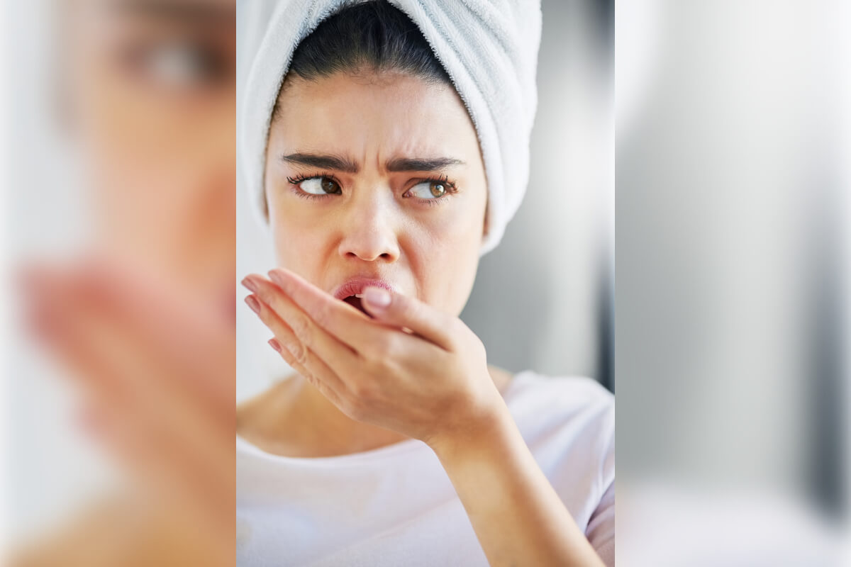 Preventing Bad Breath (Halitosis) - A woman with a concerned expression is checking her breath by cupping her hand in front of her mouth in a bathroom setting. She is wearing a white towel wrapped around her hair, indicating she might have recently showered. Her facial expression suggests worry or discomfort, likely related to her breath. The image highlights the importance of personal hygiene practices in managing and preventing bad breath (halitosis). Regular oral care, including brushing, flossing, and using mouthwash, plays a crucial role in addressing this common issue.