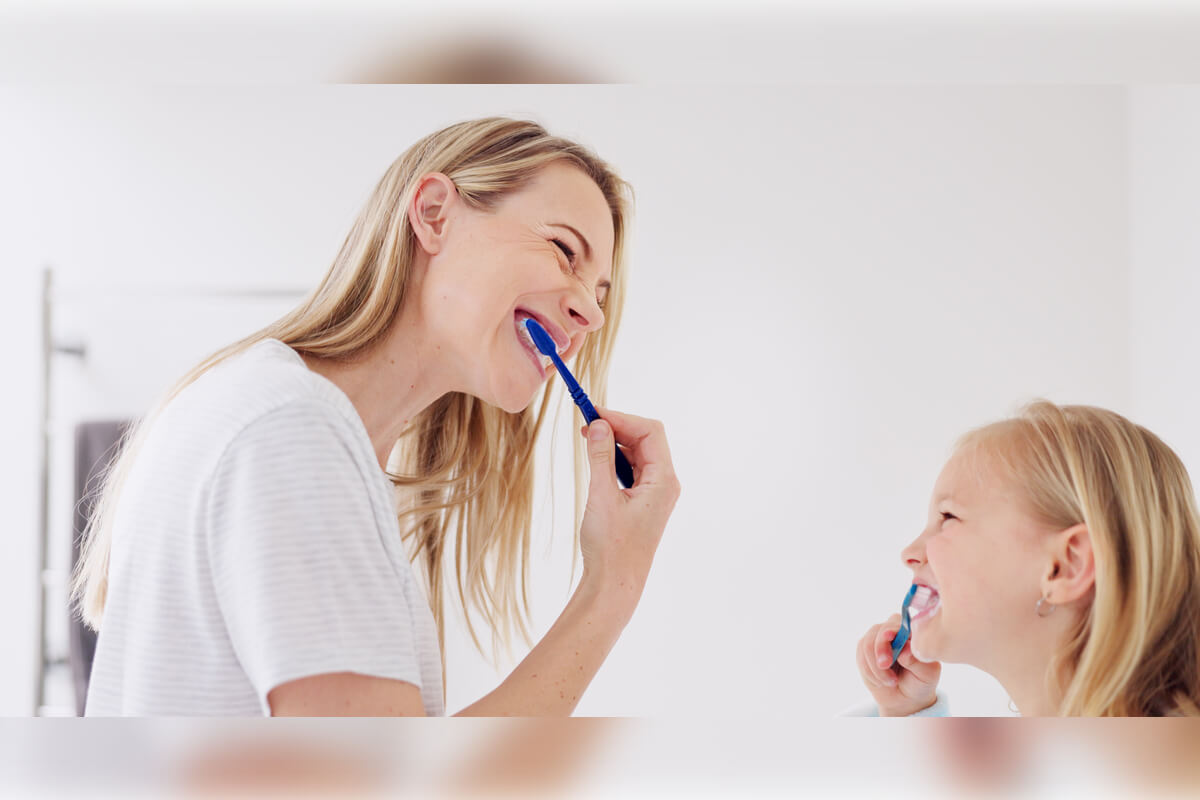 Step-by-Step Guide to Brushing Your Teeth - A mother and her young daughter are smiling as they brush their teeth together in a bright, clean bathroom. The mother, holding a blue toothbrush, is demonstrating proper brushing techniques, while the daughter enthusiastically mimics her actions with her own toothbrush. The scene emphasizes the importance of teaching children good oral hygiene habits from a young age, with the shared activity highlighting a positive, engaging approach to dental care. The light and cheerful setting reinforces the idea that brushing teeth can be a fun and essential part of a daily routine.