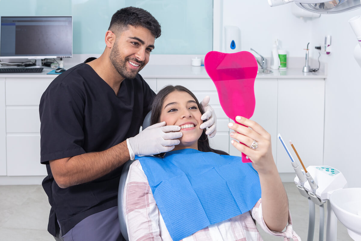 The Art of Gum Contouring - A young female patient is seated in a dental chair, smiling and looking at her reflection in a tooth-shaped pink hand mirror. A dentist, wearing a black scrubs and white gloves, is gently examining her gums and teeth, showcasing the results of a gum contouring procedure. The setting is a modern dental clinic with various dental tools and equipment in the background. This procedure is aimed at reshaping the gum line to improve the patient's smile aesthetics and oral health, demonstrating the effectiveness of advanced cosmetic dentistry techniques.