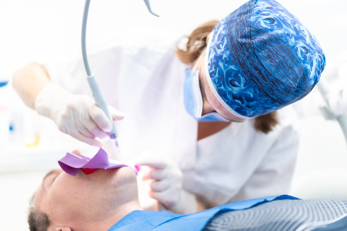 The Art of Gum Contouring - In this image, a dentist is seen performing a gum contouring procedure on a male patient, who is lying back in a dental chair. The dentist, wearing a blue patterned surgical cap, face mask, and gloves, is using a specialized dental laser tool to carefully sculpt the patient's gum tissue. A dental dam is used to isolate the area, ensuring a clean and controlled environment. The procedure, taking place in a bright and modern dental office, focuses on reshaping the gum line to reduce excess gum tissue and enhance the appearance of the patient’s teeth. This cosmetic dentistry technique aims to improve both the aesthetics of the smile and the overall oral health of the patient, demonstrating the precision and skill required in contemporary gum contouring treatments.