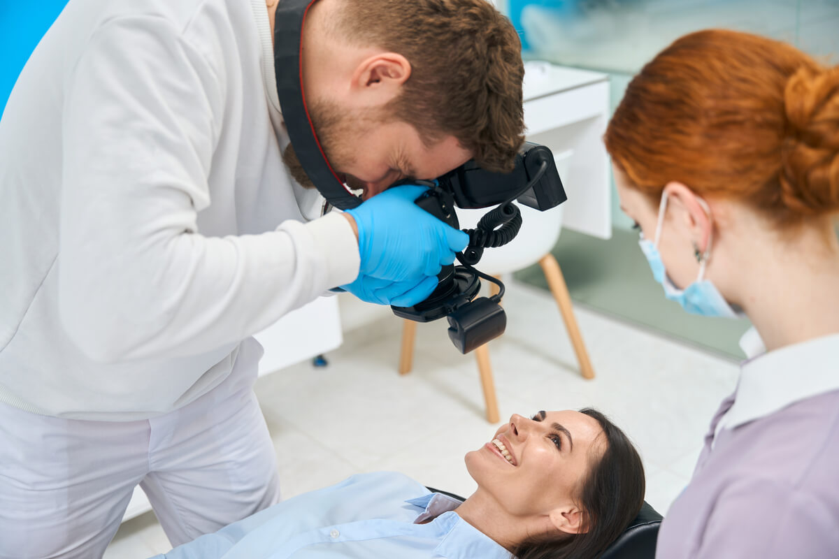 The Science of Smiling - In a modern dental clinic, a dentist uses a high-tech camera with magnification to take close-up photographs of a patient's teeth, focusing on her smile. The patient, a woman in her 30s, lies back in the dental chair, showing a relaxed and natural smile. A dental assistant, wearing a mask and observing closely, highlights the collaborative approach in dental diagnostics. This scene illustrates the role of digital imaging technology in assessing oral health, planning cosmetic enhancements, and understanding the biomechanics of smiling. It emphasizes how precision and technology are integral to achieving a perfect smile and improving patient confidence.