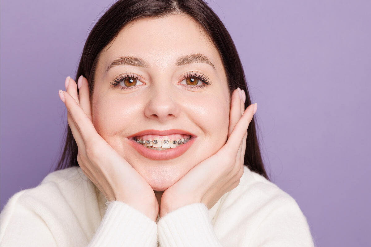 Types of Braces for Teeth - A young woman with straight, brown hair smiles confidently, showcasing her metal braces. She gently cups her face with both hands, emphasizing her braces, which are traditional metal brackets with a wire running through them. The background is a soft purple, creating a calm and inviting atmosphere. This image highlights the use of traditional metal braces, a common orthodontic treatment for aligning and straightening teeth to improve dental health and appearance.